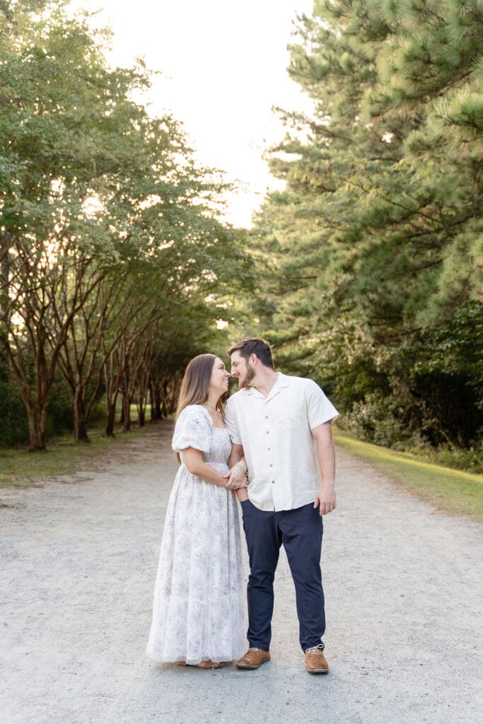 Woman wear light floral dress and man wearing white button up while posing for engagement photos surrounded by trees at Oak Grove Lake Park in Virginia