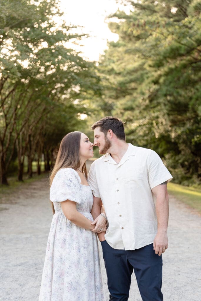 Woman wear light floral dress and man wearing white button up while posing for engagement photos surrounded by trees at Oak Grove Lake Park in Virginia